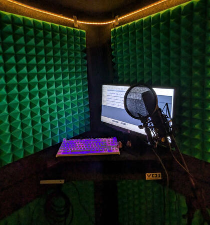 Untitled a booth constructed of multiple layers of paneling and a yellow string light. A RODE nt1 is depicted along with a monitor and keyboard.